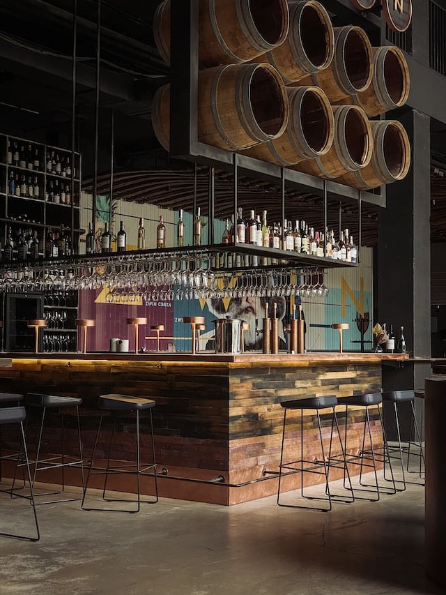 A bar with wine bottles and empty barrels above, Wine Advertising Agency.